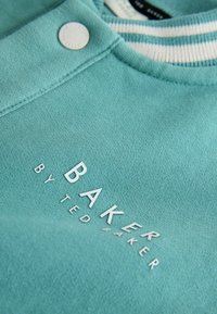 Tyrkysová mikina s rebrovaným výstrihom, ktorá má biely pruhovaný vzor a strieborné logo "BAKER by TED BAKER" na prednej strane.