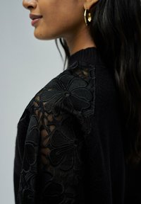 Un pull noir avec des détails en dentelle florale sur les manches et un col côtelé. Texture chaude avec une coupe décontractée.