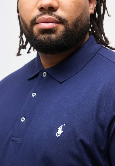 Άνθρωπος με γενειάδα που φοράει μπλε μανόλο πόλο με λευκό κεντητό λογότυπο παίκτη πόλο στο στήθος, το γιακά κλειστό με κουμπί.