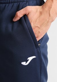 Marineblå atletiske bukser med en glat overflade, udstyret med en sidelomme og et hvidt logo på venstre lår. Elastisk talje inkluderet.