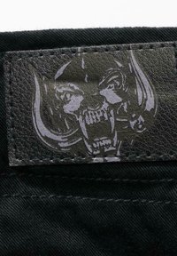 Svart teksturert stoff med et stort grått lærpatch med et hodeskalle-design og hoggtenner, sydd langs kanten.