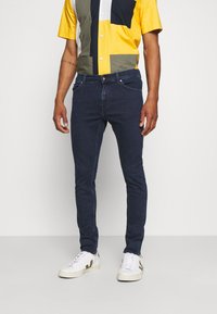 Vaqueros de mezclilla oscura con un corte ajustado, combinados con una camisa de mangas cortas multicolor que presenta secciones amarillas, azul marino y olivo. Zapatillas blancas.