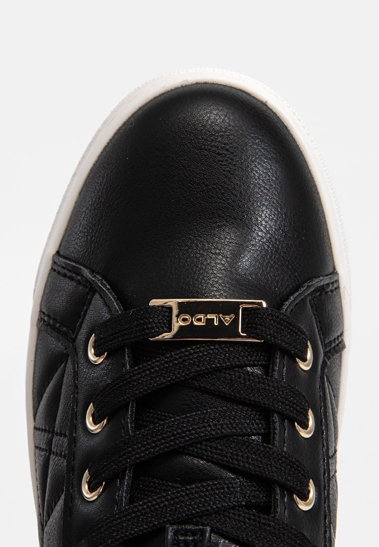 Svarta lädersneakers med svarta snören trädde genom guldögon och en guld varumärkesplåt på toppen, mot en vit bakgrund.