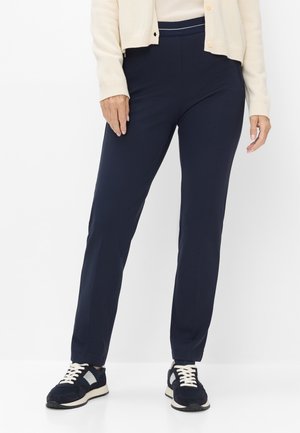 Frau trägt dunkelblaue Hose, schwarz-weiße Sneakers und eine cremefarbene Strickjacke vor einem schlichten weißen Hintergrund.