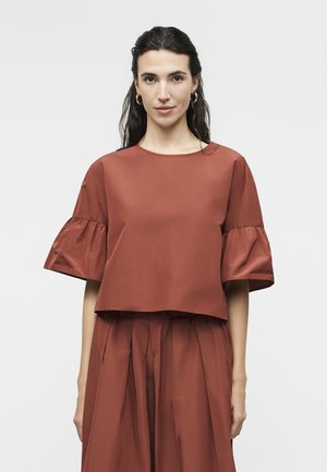 Γυναίκα με σκούρα μαλλιά που φοράει μπλούζα κοντομάνικη σε σκουριά απόχρωση με βολάν στα μανίκια και ταιριαστό πλισέ φούστα, κοιτάζοντας μπροστά πάνω σε απλό φόντο.