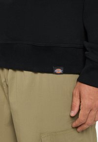 Black knit sweatshirt with ribbed cuffs and hem. Logo patch on the bottom. Worn with khaki cargo pants showing pocket detail.