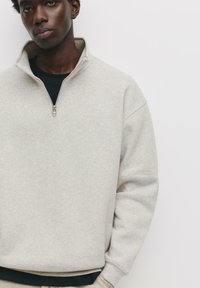 Pulover din fleece gri deschis, cu guler cu fermoar pe un sfert, cu manșete și tiv din material rib elastic. Este purtat peste o cămașă neagră, având o croială relaxată.