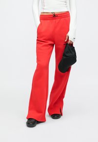 Κόκκινο φλις παντελόνι φόρμας με φαρδύ πόδι, ελαστική μέση και κορδόνι, με πλαϊνές τσέπες και σκισίματα στα τελειώματα. Μαύρο καπέλο στο χέρι.