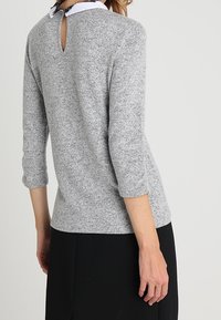 Grå stickad topp med trekvartsärmar, med ett nyckelhål baktill och lagerkrage över en mörk kjol. Mjuk textur, avslappnad design.