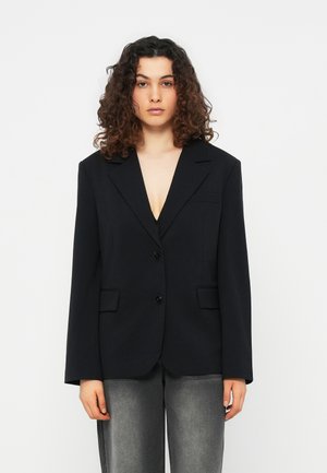 Γυναίκα με μπουκλωτά μαλλιά, που φοράει μαύρο oversized σακάκι και ξεθωριασμένο γκρι τζιν, στέκεται μπροστά σε απλό λευκό φόντο.