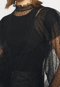 Schwarze, transparente Bluse mit Punkten, Rüschenausschnitt, gefütterten Details und strukturierten Ärmeln. Der Stoff wirkt leicht und zart.