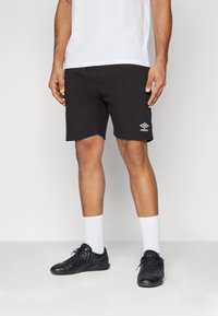 Pantalones cortos deportivos negros hechos de una tela suave, con un logo bordado en blanco. Combinados con calcetines blancos y zapatillas deportivas negras.