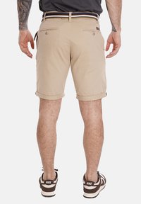 Pantaloni corti color khaki con risvolti, due tasche posteriori con bottoni e una cintura a righe. Indossati con sneakers scure con dettagli bianchi.