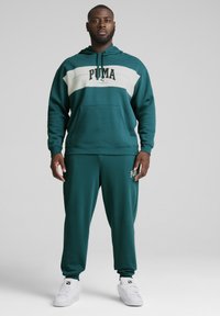 Felpa di un verde scuro con pantaloni jogger abbinati, caratterizzati da una striscia orizzontale bianca e un logo. Presenta un cappuccio e una tasca frontale; il tessuto ha una texture liscia.