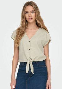 Blusa verde chiaro a maniche corte con scollo a V, chiusura con bottoni frontale e orlo annodato. Abbinata a una gonna in denim scuro. Tessuto dalla consistenza liscia.