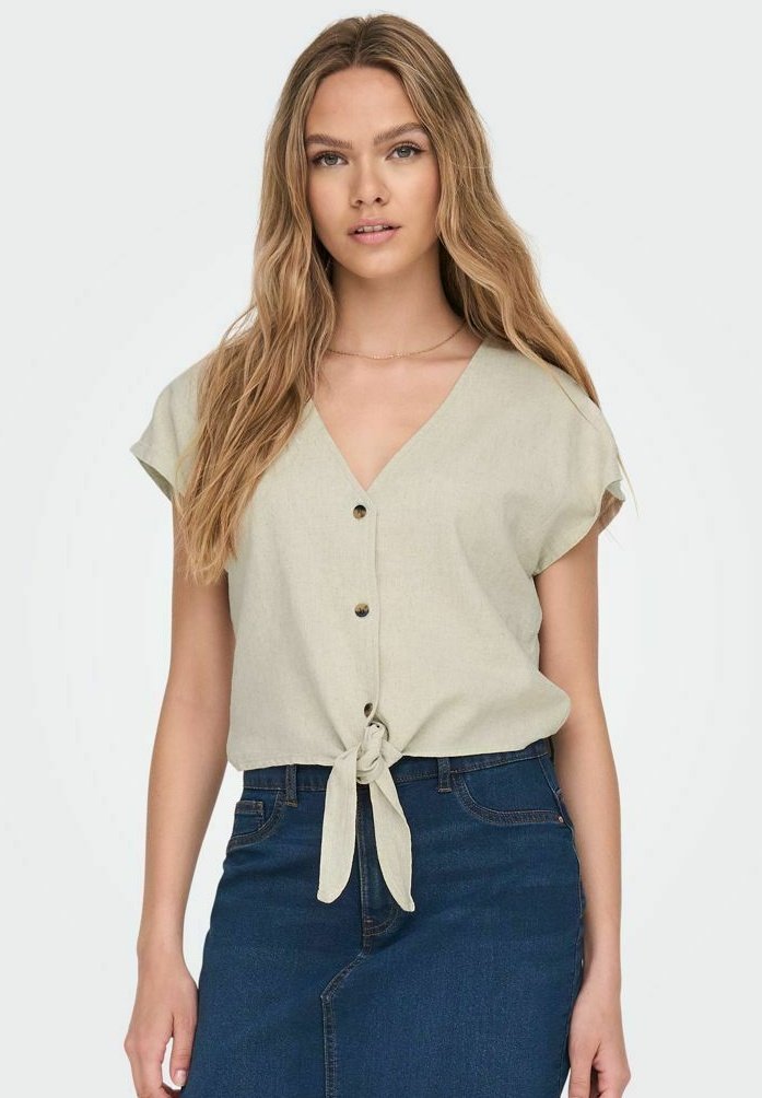 Blusa verde chiaro a maniche corte con scollo a V, chiusura con bottoni frontale e orlo annodato. Abbinata a una gonna in denim scuro. Tessuto dalla consistenza liscia.