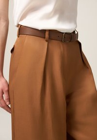 Pantalones marrones con una textura suave y ligeros pliegues, combinados con un cinturón de cuero marrón que presenta una hebilla metálica rectangular.