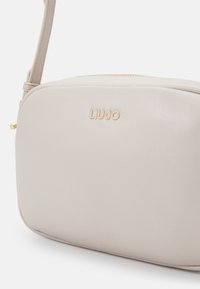 Béžová kožená crossbody taška s obdélníkovým tvarem, zlatým logem "LIUJO" a zapínáním na zip. Obsahuje úzký nastavitelný popruh.