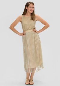 Robe plissée longueur genou en or avec un effet scintillant. Design sans manches et taille cintrée. Associée à des sandales plates.