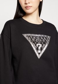 Sudadera negra con un diseño triangular de pedrería plateada con un patrón de diamantes y texto. Textura suave, ajuste holgado, escote amplio.