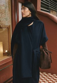 Woman wearing a dark navy belted cape and scarf, holding a brown suede bag, standing in tiled hallway with patterned wall and window.
