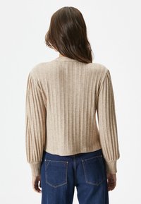 Beige gerippter Pullover mit langen, puffigen Ärmeln und einem cropped Schnitt, kombiniert mit blauen Jeans, die sichtbare Hintertaschen haben.