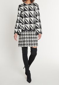 Šaty s geometrickým vzorem v černobílé barvě, dlouhými rukávy, kulatým výstřihem a přiléhavým střihem, doplněné vysokými černými botami.