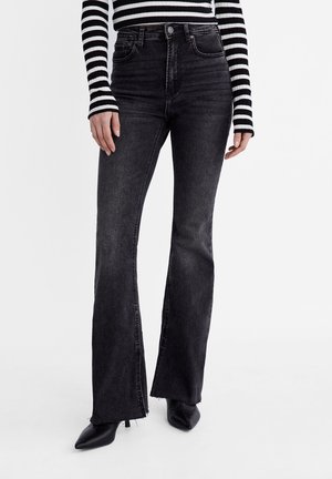 Person iført sorte højtaljede flare-jeans med rå kanter, kombineret med sorte spidse hæle og en sort-hvid stribet langærmet top.