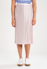 Kvinna som bär en metallisk rosa plisserad midikjol, en blå och vit randig topp samt vita sneakers, stående mot en enfärgad bakgrund.