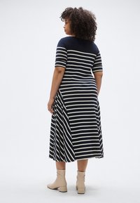 Tengerészkék és krémszínű csíkos ruha, rövid ujjú, laza szabású, térdig érő, oldalsó sliccsel. Bézs bokacsizmával viselve.