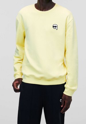 Mężczyzna ubrany w jasnożółtą bluzę z małym czarno-białym karykaturalnym logo na piersi oraz czarne spodnie z dopasowanym logo.