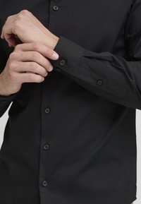 Zwarte button-up shirt van gladde stof, met lange mouwen, opgerolde manchetten en zwarte knoopsluitingen aan de voorkant.