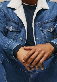 Jeansjacka med en vit, sherpa-fodrad krage, som har två bröstfickor och silverdetaljer. Bärs över en mörk marinblå stickad tröja.