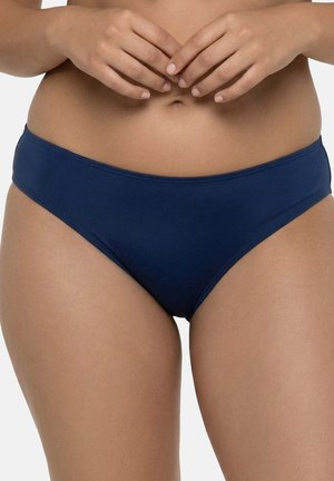 Kvinde iført ensfarvede mørkeblå bikinibunddele med hænderne let foldet over navlen, neutral baggrund.