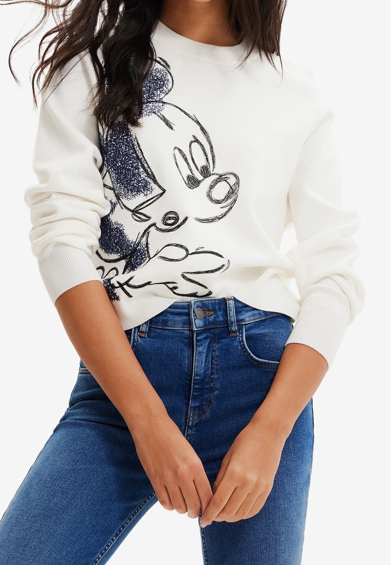 Femme portant un sweat-shirt blanc avec un motif Mickey Mouse style croquis et un jean en denim bleu, mains légèrement jointes devant elle.