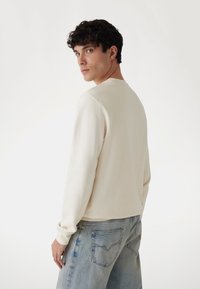 Felpa a maniche lunghe color crema con scollatura a girocollo, realizzata in tessuto morbido. Abbinata a jeans in denim blu chiaro con tasche posteriori cucite.