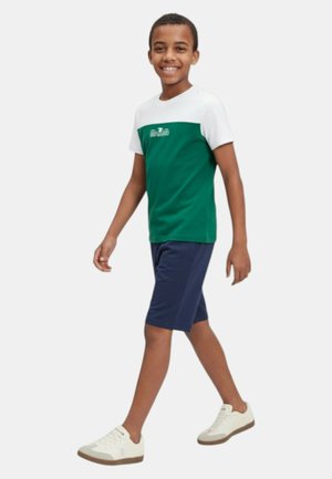 Ragazzo che cammina, indossa una maglietta a maniche corte verde e bianca, pantaloncini blu navy e sneakers bianche, sorride, su uno sfondo chiaro e uniforme.