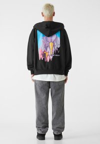 Sort hættejakke med et farverigt grafisk design, der har pastelnuancer og teksten "LOST YOUTH™" på ryggen, båret over grå jeans.