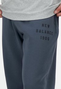 Pantalones de chándal gris oscuro, fabricados en tejido suave, con un logo impreso en negro "NEW BALANCE 1906" en el muslo izquierdo. Cintura elástica.