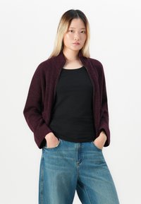 Une jeune femme aux longs cheveux blonds porte un sweat à capuche zippé violet foncé par-dessus un haut noir, associé à un jean bleu, les mains dans les poches.