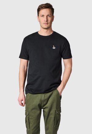 Man draagt een zwart t-shirt met klein duikerslogo en olijfgroene cargobroek, staand met één hand in de zak tegen een effen achtergrond.