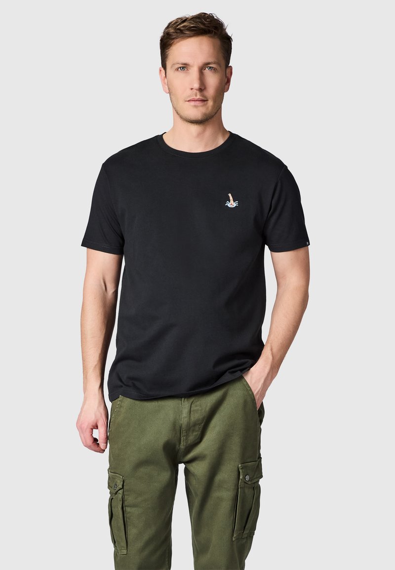 Mann trägt schwarzes T-Shirt mit kleinem Taucher-Logo und olivgrüner Cargohose, steht mit einer Hand in der Tasche vor schlichtem Hintergrund.
