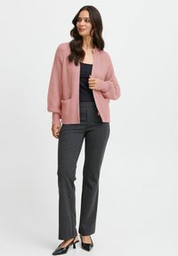 Rosa stickad cardigan med långa ärmar, framfickor, bärs över en svart topp, tillsammans med grå byxor och svarta spetsiga skor.