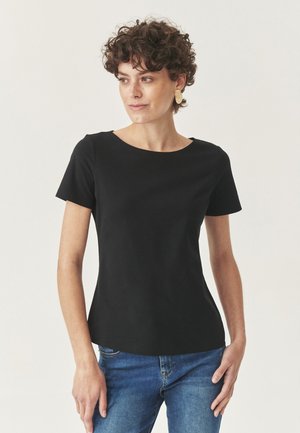 Femme aux cheveux courts et bouclés portant des boucles d'oreilles en or, un haut noir uni à manches courtes et un jean bleu, se tenant devant un fond clair.