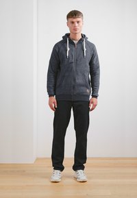 Siva zip-up majica s kapuljačom, prednjim džepovima i rebrastim manžetama, u kombinaciji s crnim hlačama i bijelim cipelama na drvenom podu.