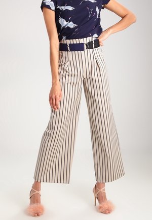 Femme portant un pantalon large beige à rayures verticales noires, une ceinture bleu marine, un haut bleu marine imprimé de grues blanches, et des talons hauts roses duveteux.