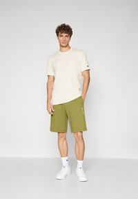 Hellbeige T-Shirt mit "Champion"-Logo, kombiniert mit grünen Shorts. Weiße Sneaker und Socken mit sichtbarem Logo. Einfaches und lässiges Design.