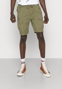 Shorts cargo vert olive avec plusieurs poches, taille à cordon de serrage et coupe décontractée. Portés avec des baskets montantes blanches et brunes.