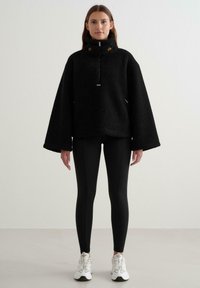 Fekete bolyhos pulóver magas gallérral, széles ujjal és cipzáros záródással. Fekete leggings-szel és fehér sportcipővel párosítva.