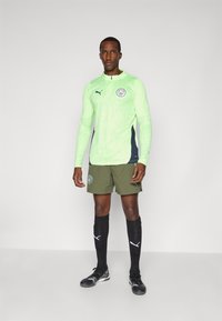 Helle grüne langärmelige Sportshirt mit marineblauen Akzenten und Logo, kombiniert mit olivgrünen Shorts und schwarzen Socken, die weiße Logodetails aufweisen.